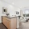 A modern kitchen with a spacious island and light wood cabinetry, seamlessly flowing into a sunlit living area with access to a private balcony.